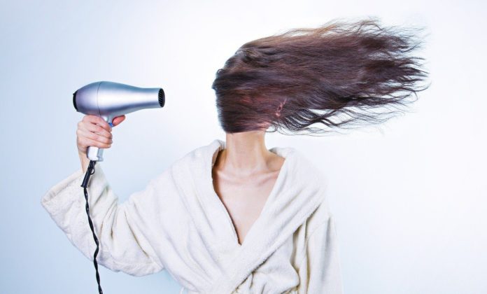 UNA DONNA IN ACCAPPATOIO TIENE UN ASCIUGACAPELLI NELLA MANO DESTRA CHE LE FA VOLARE I CAPELLI VERSO SINISTRA COPRENDOLE IL VOLTO