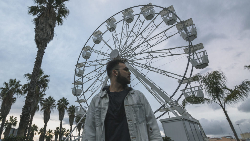 Damiano Mulino, in occasione del nuovo singolo dal titolo Come l'America, posa su uno sfondo con le palme e una ruota panoramica, in giacca di jeans e maglietta nera, con lo sguardo rivolto verso il lato