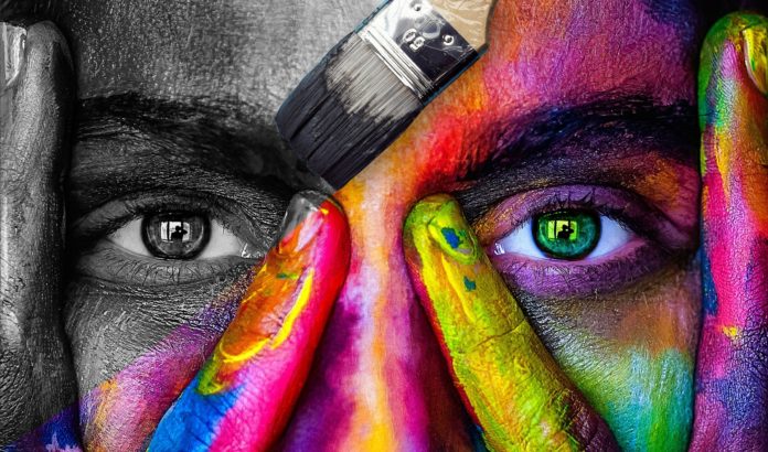 Arte Fiera Bologna 2020 una faccia di donna completamente dipinta con tanti colori, un pennello che sta dipingendo in bianco e nero l'occhi sinistro e nela pupilla il riflesso di un fotografo
