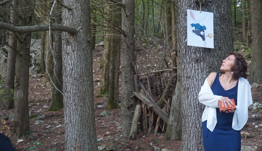 Giulia Ventisette interpreta Alice nel paese delle cianfrusaglie, indossa un vestito attillato blu lungo, con un coprispalle bianco, in mezzo a un bosco, tiene una scatoletta marrone in entrambe le mani, mentre si gira a guardare l'immagine del bianconiglio pinzato su un albero