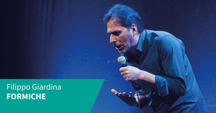 Filippo Giardina con camicia blu e microfono in mano di profilo parla sull'angolo basso a sinistra il suo nome e quello del suo show, 