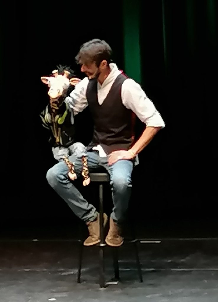 Al Super Fantasy Magic Show, il ventriloquo Rafael Voltan, seduto su uno sgabello, vestito con jeans, camicia bianca e gilet nero, tiene in braccio la giraffina Gi, che ha gli occhi puntati verso il pubblico