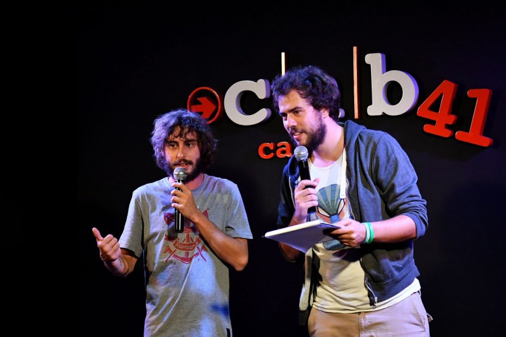 i panpers in uno sketch al cab 41, mentre tengono un microfono ciascuno in mano, stanno parlando con il pubblico, uno dei due personaggi a sinistra indossa una maglietta a maniche corte grigia con un disegno arancione in mezzo, a destra invece l'altro indossa pantaloni color crema, una maglietta bianca con un disegno in mezzo e una felpa blu, su sfondo nero con la scritta dietro del locale, cab 41, cabaret