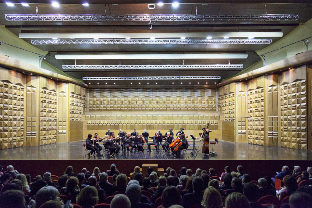 sala dell'auditorium conciliazione di roma dedicata ai concerti, dotata di un grande palco che ospita un'orchestra, con il pubblico che sta partecipando al concerto, dietro ci sono le pareti del palcoscenico decorate a quadrati tridimensionali gialli, color crema e bianchi, su in alto ci sono i riflettori