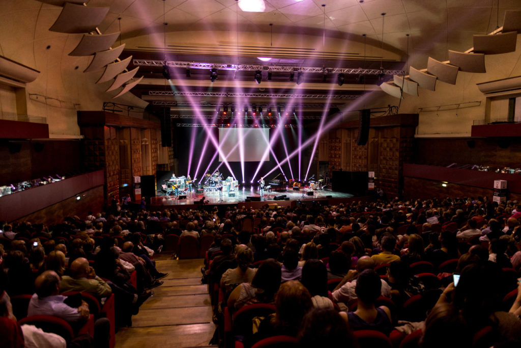 palcoscenico dell'auditorium conciliazione di roma, in scena c'è un concerto al quale stanno partecipando numerosi spettatori, seduti nelle poltrone della sala dell'auditorium, le luci sono soffuse in tutta la sala, e dal palcoscenico partono dei riflettori in diverse direzioni verso l'alto di colore rosa