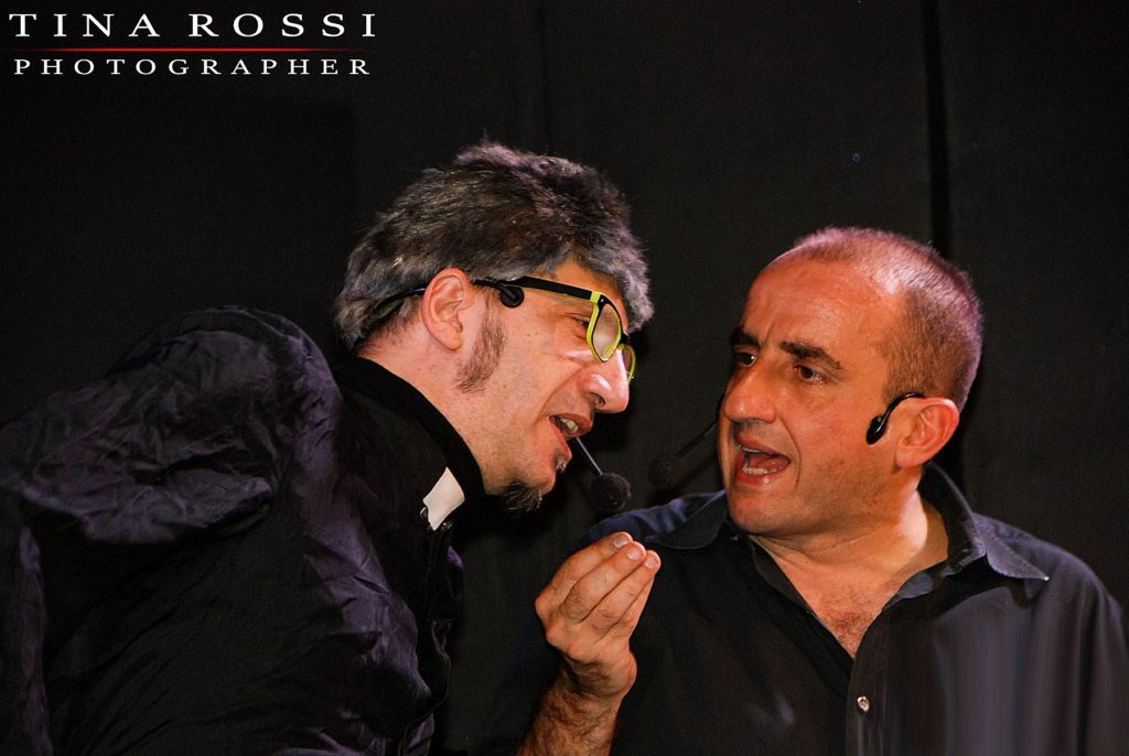 Mauro Villata e Gianpiero Perone in scena durante uno sketch, a sinistra Villata, nelle vesti da frate, indossa un paio di occhiali da vista, a destra Perone con una mano alzata all'altezza del mento in mezzo ai due sta parlando con il comico, indossa una camicia nera, su sfondo nero, fotografia di Tina Rossi