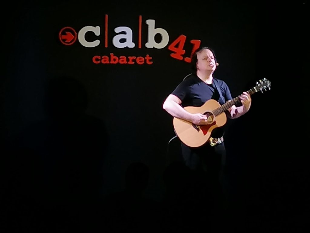 Dado, comico di Zelig e Colorado, su sfondo nero con il nome del locale Cab41, cabaret, suona la chitarra mentre sta cantando, indossa una maglietta a maniche corte nera