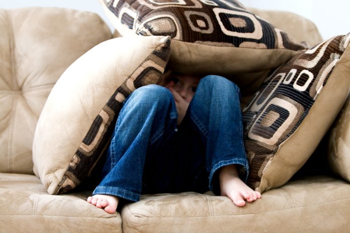 fobia, paura, che un bambino dimostra, nascondendosi tra un ammasso di cuscini sul divano. Indossa i jeans ed è scalzo. I cuscini e il divano sono color crema, e i cuscini hanno un pattern a quadrati di varie sfumature di marrone e bianco