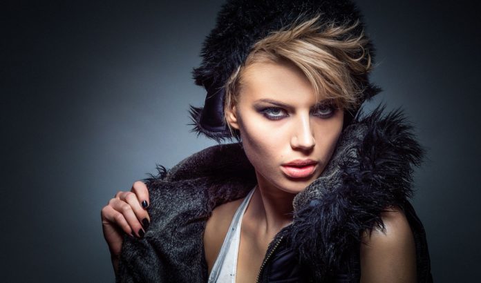 modella fashion style in primo piano, con sguardo serio, bionda con un ciuffo di capelli corti da un lato, indossa un cappello e un gilet nero, su sfondo di colore grigio scuro