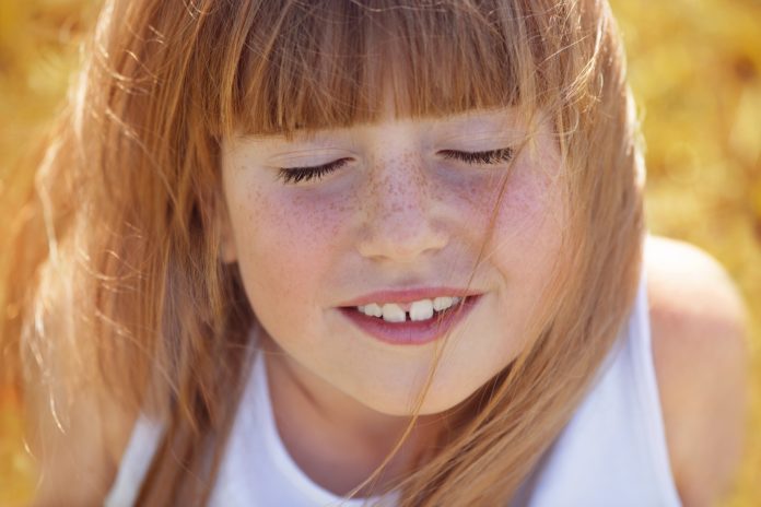 una bambina con le lentiggini, i capelli castano chiari e la frangia, tiene gli occhi chiusi e sorride mostrando i de tni, con i capelli mossi dal vento, indossa una canotta bianca, sfondo arancione