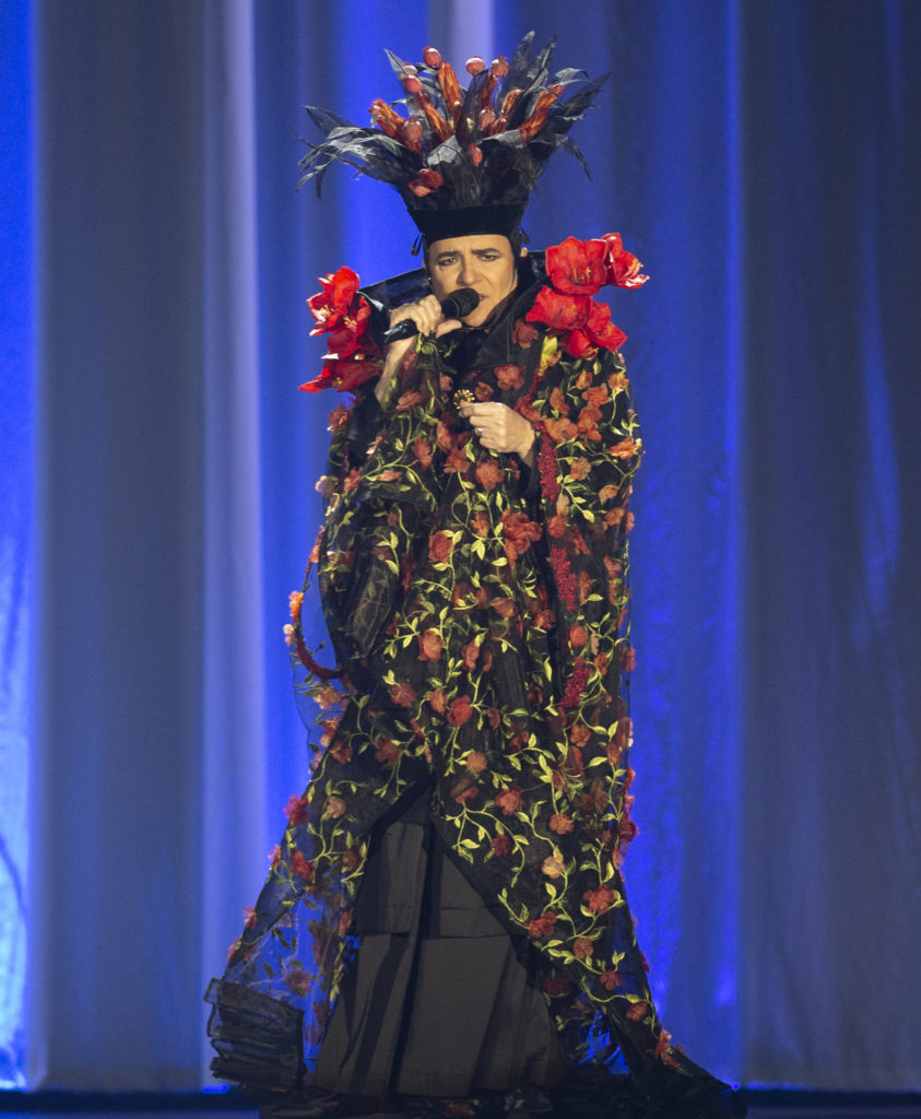 Renato Zero con microfono in mano canta con un vestito lungo colorato con tanti fiori e indossa un cappello vistoso con delle rose