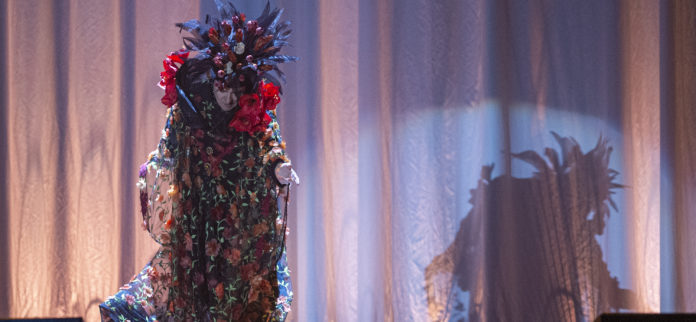 Renato Zero nel tour di Zero il Folle veste un abito largo con fiori e indossa un cappello con delle rose, è inchinato mentre ringrazia il pubblico