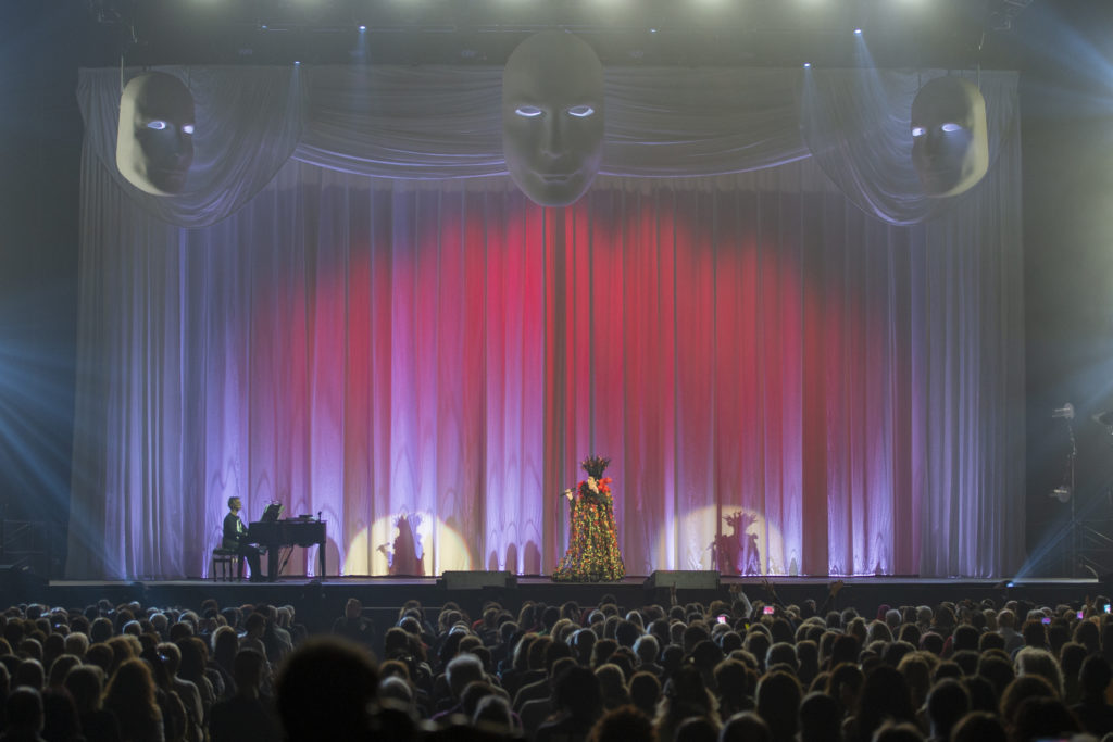 il concerto del tour di Zero il folle comincia con il sipario chiuso. stefano Senesi seduto al piano forte e Renato con un vestito largo a fiori e un cappello vistoso canta