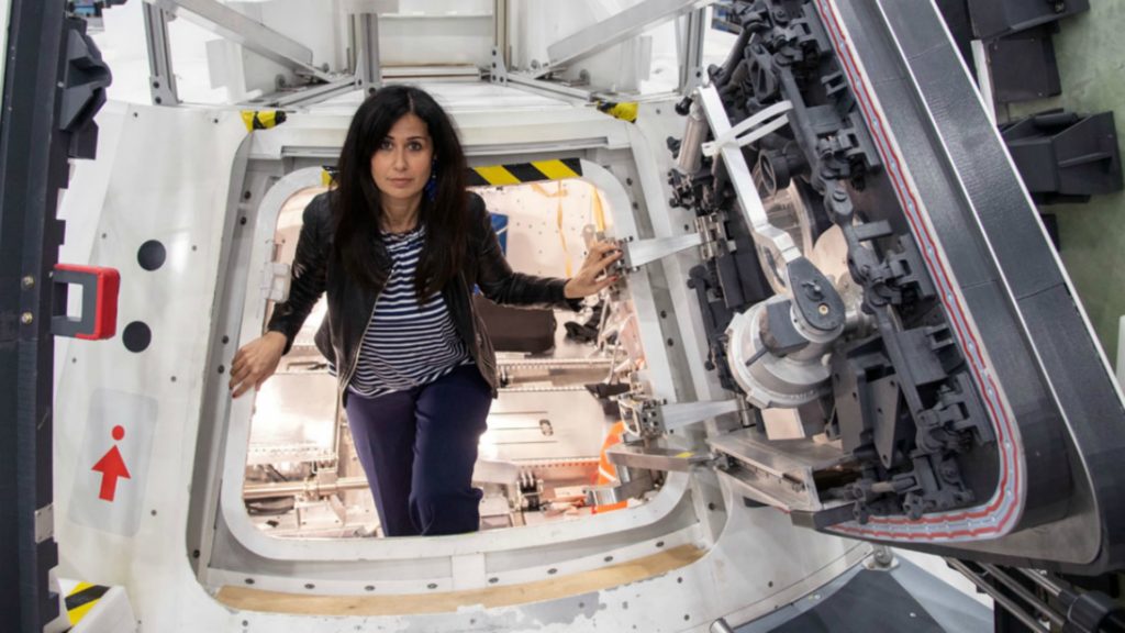 alessandra bonavina all'interno di una capsula spaziale, in mezzo a una porta quadrata piccola della capsula, appoggia le mani alle pareti, indossa una maglietta a righe bianche e blu, una giacca in pelle nera e un paio di pantaloni blu, ha i capelli sciolti neri e lunghi fino alle spalle e lo sguardo con un accenno di sorriso