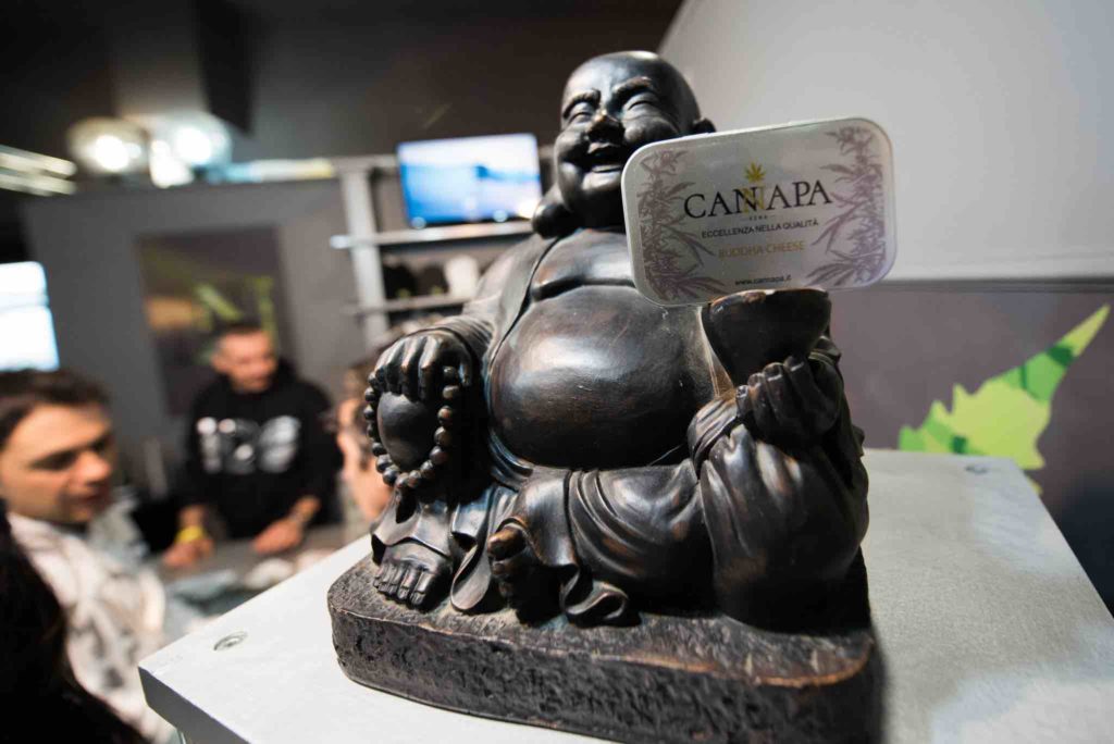 una statuetta del budda in nero che tiene in una mano un cartoncino con su scritto canapa, dietro ci sono alcune persone della fiera