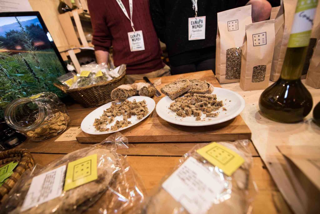 su un tavolo di uno stand della fiera canapa mundi dell'anno scorso ci sono due piatti con delle fettine di pane a base di semi di canapa, dietro il bancone ci sono due espositori vestiti uno con un maglioncino bordeaux e l'altro in nero, sul tavolo ci sono anche dei prodotti confezionati con sopra un'etichetta che indica la ricetta e gli ingredienti