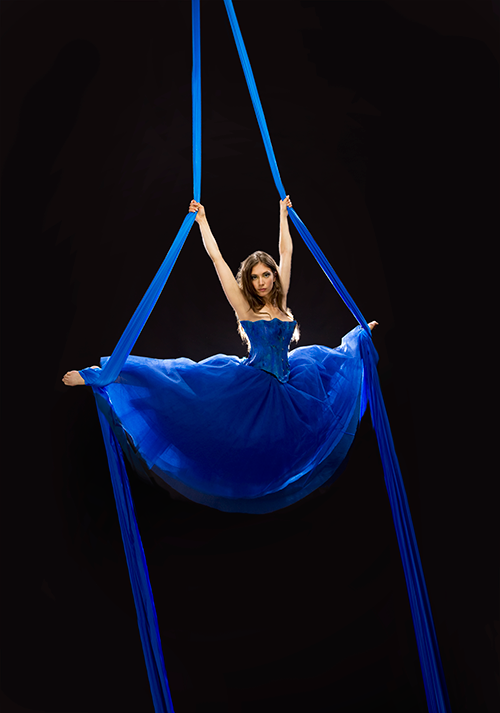 erika lemay, su sfondo nero, in un'esibizione l'artista si regge con le braccia e i piedi in alto a due corde di tessuto blu, in una spaccata, indossa una vestito senza spalline lungo e blu, ha i capelli castani e sciolti sulle spalle, con sguardo sensuale
