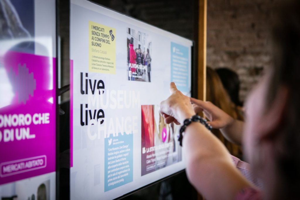 due mani che indicano una vetrata sulla quale c'è un poster relativo del progetto live museum live change