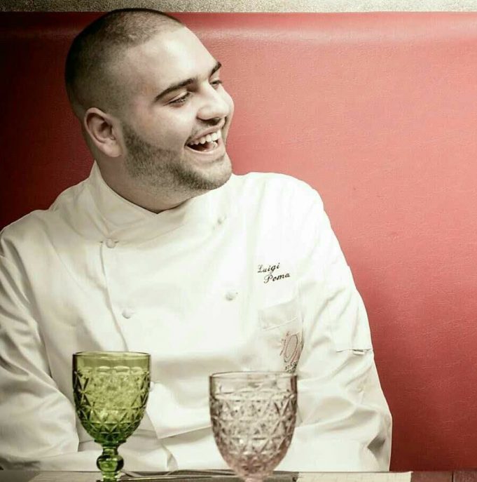 Paolo Palumbo in veste da chef, sorride mostrando i denti, quando non aveva ancora la SLA, davanti a lui su un tavolo ci sono due bicchieri di vetro, uno verde e uno trasparente, lo sfondo è rosso