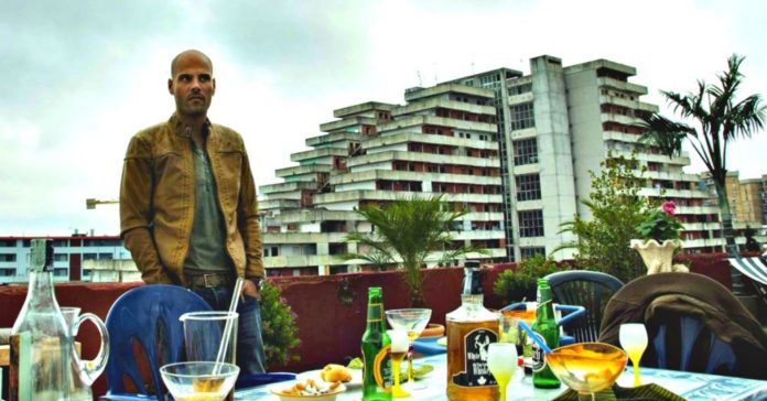 Vele, Scampia, Gomorra. Ciro L'Immortale, protagonista di Gomorra ha davanti a sè un tavolo con bicchiere e bottiglie di alcool. Alle spalle l'edificio simbolo di Scampia: le vele.