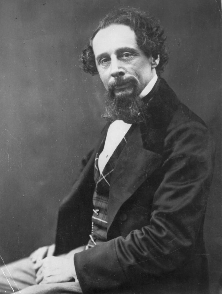 charles dickens, ritratto in bianco e nero, seduto rivolto verso sinistra, appoggia le braccia sulle gambe, con la testa leggermente inclinata, accenna un sorriso, su sfondo nero 