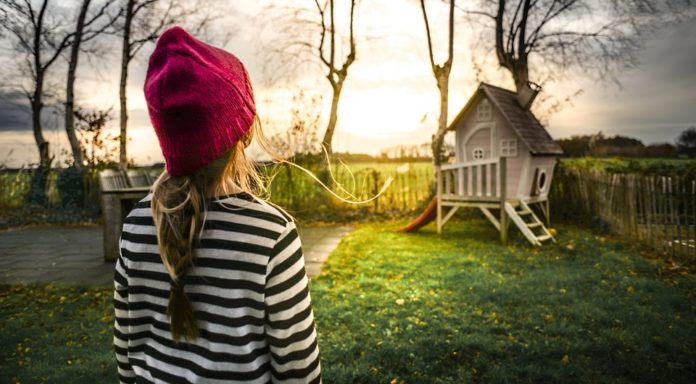 una bambina con i capelli lunghi e raccolti in una treccia, girata di schiena, indossa una maglietta a maniche lunghe a righe bianche e nere e un cappellino rosso, guarda in lontananza una piccola casetta di legno, in un cortile, dietro la casetta ci sono quattro alberi molto alti e spogli, con il tramonto