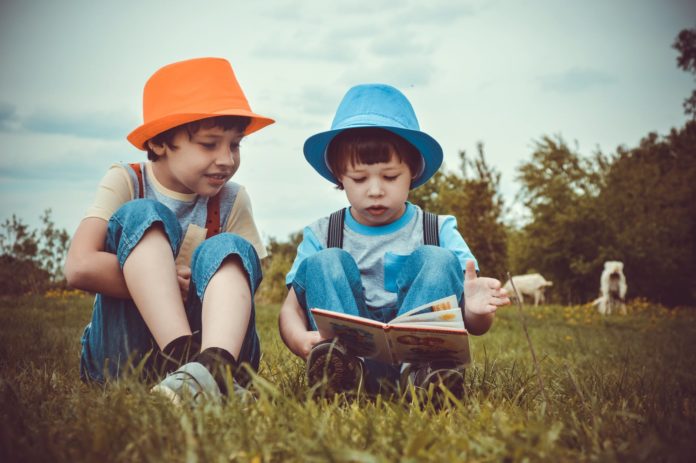 per il premio nazionale nati per leggere, due bambini immersi nella lettura di un libro, due bambini piccoli che indossano un cappellino a cilindro, sono vestiti entrambi con la salopettes in jeans e maglietta a maniche corte, uno dei due bambini tiene in mano un libro, sono seduti su un prato verde, dietro in lontananza ci sono alcuni alberi e arbusti