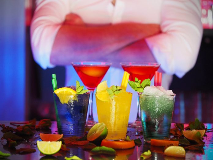 per speakeasy di the young pope, tre cocktail di diversi colori, su un tavolo di un bar con alcuni pezzi di frutta ed altri ingredienti intorno, dietro un uomo in camicia bianca con le braccia conserte, non si vede il viso, solo il busto