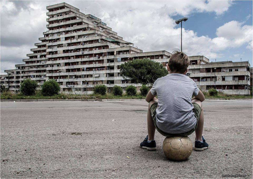 Scampia, Gomorra,vele. Bambino di schina, seduto su un pallone, guarda le vele di Scampia.