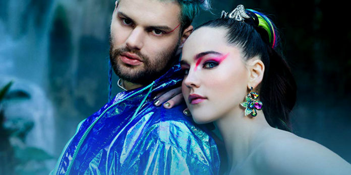 sofi tukker, per il singolo good time girl, il ragazzo ha uno sguardo serio, indossa una giacca impermeabile blu lucente, la ragazza si appoggia alla sua spalla con lo sguardo serio, indossa un paio di orecchini molto grandi, ha le spalle scoperte, entrambi sono truccati, lui ha un filo di trucco rosso che parte dalla coda dell'occhio, e lei ha un filo di trucco rosa, e ha i capelli raccolti in uno chignon