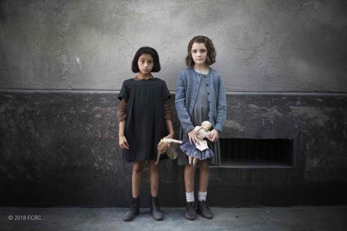 Amica Geniale, romanzo. Le due protagoniste, Lila e Lenù in piedi su un marciapiede. Entrambe le bambine reggono in mano una bambola di pezza.