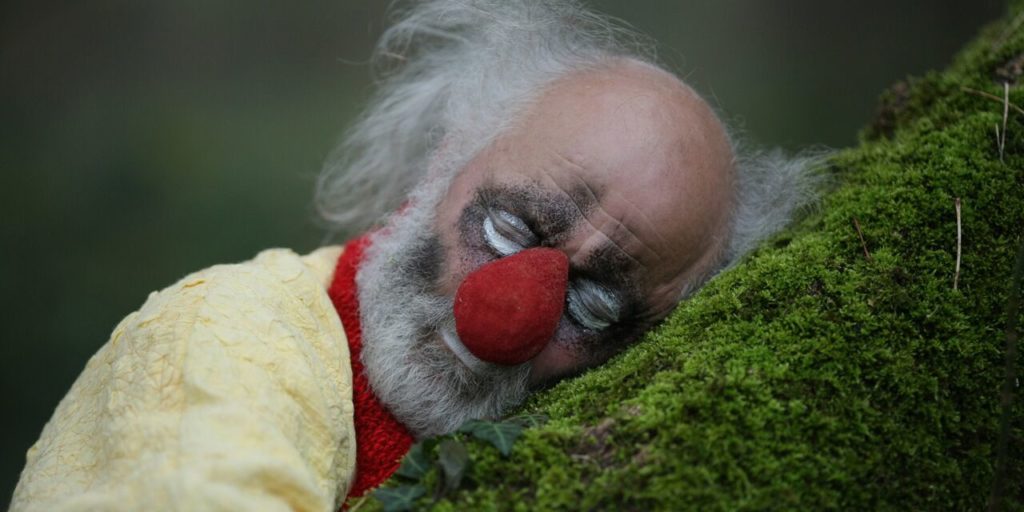 slava polunin nelle vesti da clown con un enorme naso rosso, appoggia la testa al tronco di un albero con il muschio
