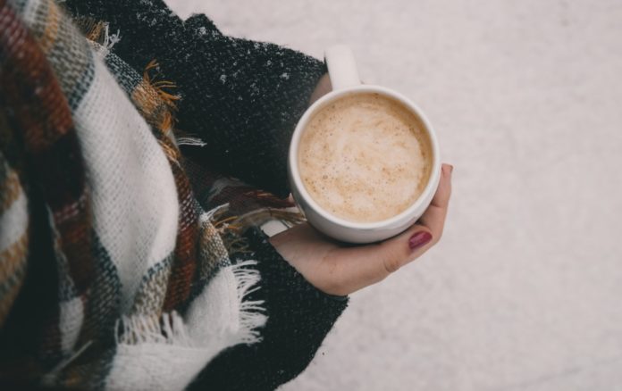 al freddo, una ragazza che indossa una giacca nera e una sciarpa bianca a quadri marroni e neri, tiene tra le mani una tazza con una bevanda calda, intorno c'è la neve, non si vede il viso della ragazza, solo dall'alto la braccia che tengono la tazza