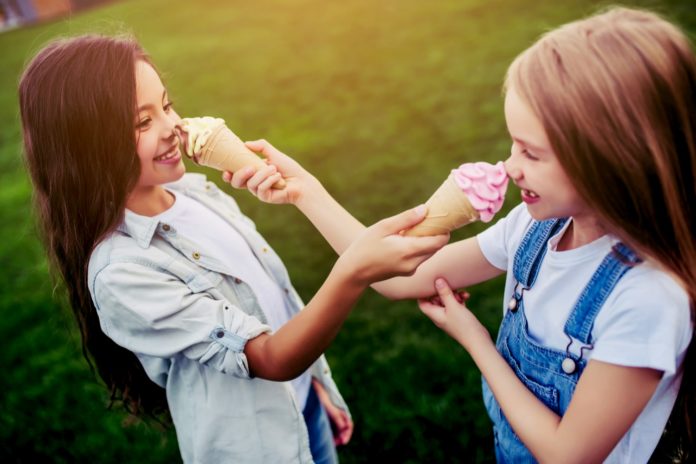 due bambine si buttano in faccia a vicenda un cono di gelato sorridendo, su un prato verde, una è vestita con una lunga camicia azzurro chiaro e jeans, e l'altra indossa una maglietta a maniche corte bianca con la salopette in jeans