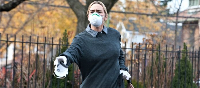 Contagion, Coronavirus. La donna, protagonista del film Contagion, indossa una camicia e un maglioncino blu. Ha la bocca coperta da una mascherina. Tiene nella mano sinistra uno zaino e con la mano destra porge, dinanzi a lei, una mascherina.