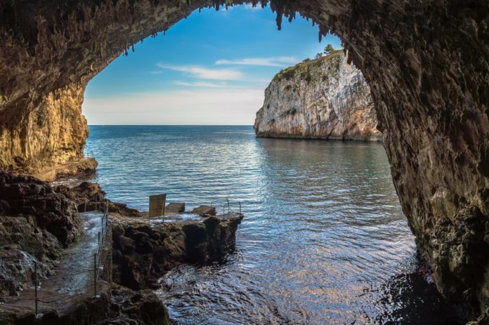 Grotta Zinzulusa, stalattiti, stalagmiti. Grotta che si affaccia sul mare di colore azzurro chiaro e limpido. Il cielo è azzurro con qualche nuvola.