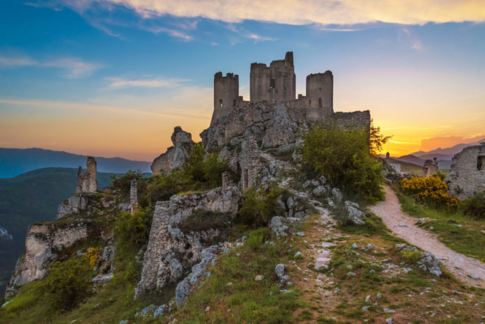 Rocca Calascio, Parco Nazionale, rocca. In primo piano il maestoso castello costruito interamente in pietra bianca. Il cielo è azzurro, con qualche nuvola bianca e il sole che tramonta.