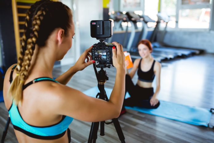 Da influencer a professionisti del fitness nella foto una ragazza con treccia bionda riprende con una telecamera un'influencer in tuta da ginnastica, seduta per terra mentre mostra dei prodotti per stare in forma