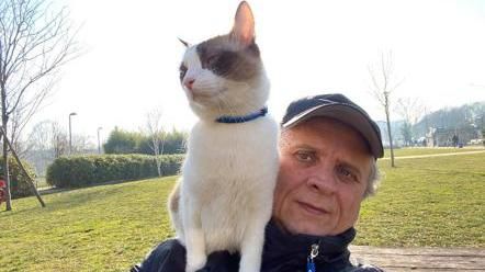 Un signore anziano  con il berretto e una tuta porta a spasso sulle spalle il suo gatto bianco con un a chiazza beige sull'occhio sinistro