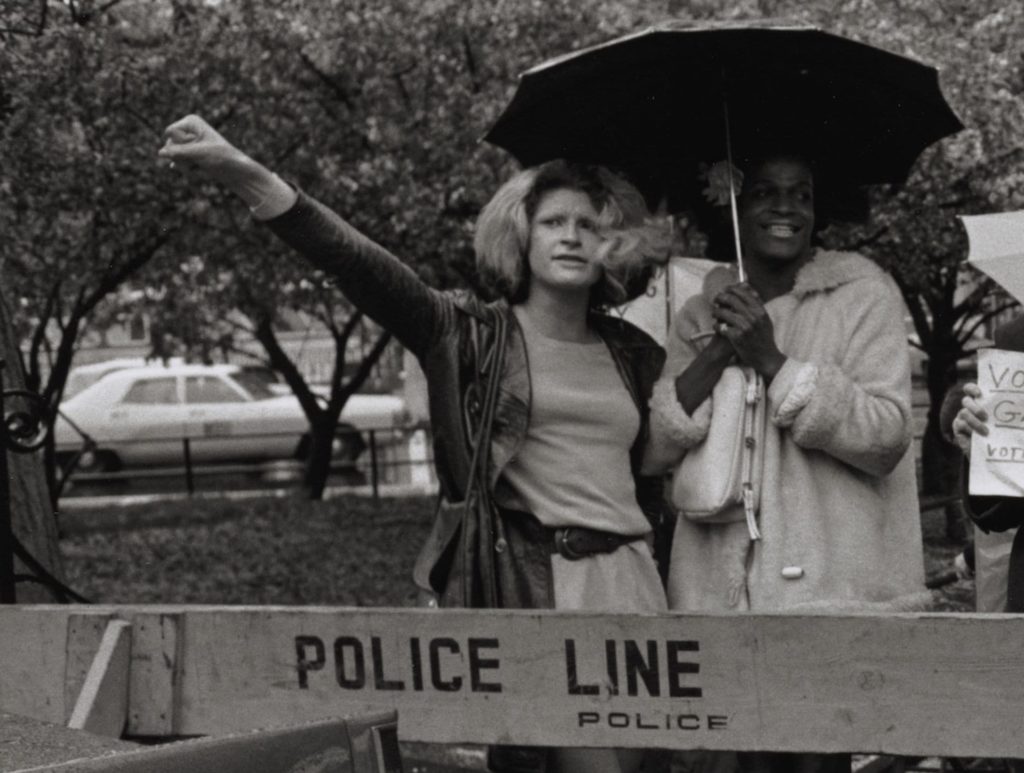 caso Marsha P Johnson nella foto in bianco e nero Marsha e Sylvia insieme sotto l'ombrello dietro a uno nastro con la scritta police line