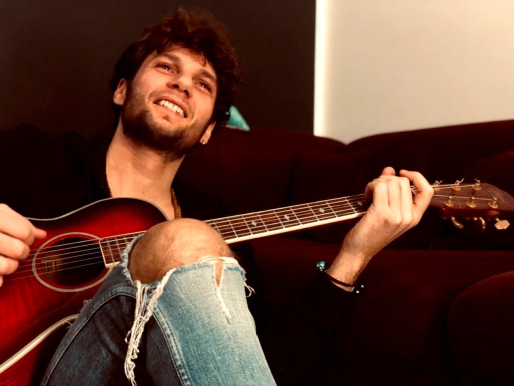 Napo un milione di visualizzazioni per "Supereroe". Nella foto il cantante, seduto e con la chitarra acustica in mano, che indossa jeans strappati, e una camicia nera. Sorride all'obiettivo.