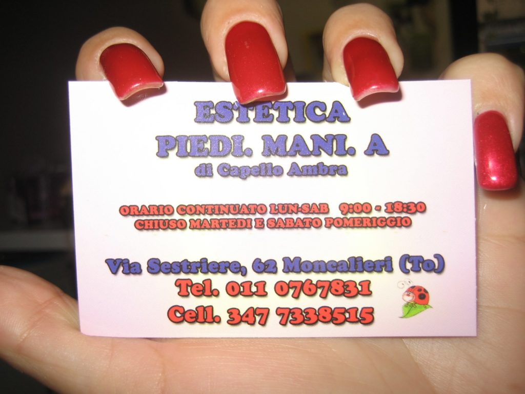 Una mano con unghie lunghe rosse tiene un biglietto da visita del centro PiediMani.A di Ambra Capello