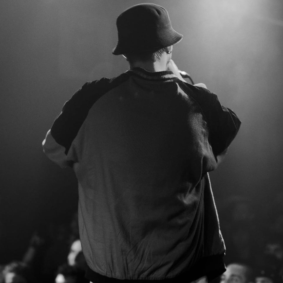 Penny - tutto di personale. Nella foto in bianco e nero, il rapper ripreso di spalle durante un concerto, che indossa un cappello nero e una t-shiert bianca con le maniche nere.