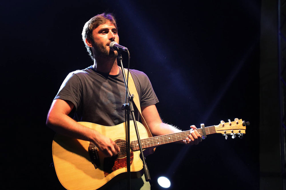 Vincenzo Capua, presenta "Weekend". Nella foto il cantautore romano, durante una esibizione, davanti al microfono, che imbraccia la chitarra acustica, e indossa una t-shirt marrone