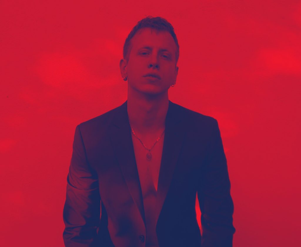 Giacomo EVA "tropici". nella foto il giovaane cantante, dietro uno sfondo rosso, che indossa una giacca nera, petto nudo e catenina al collo