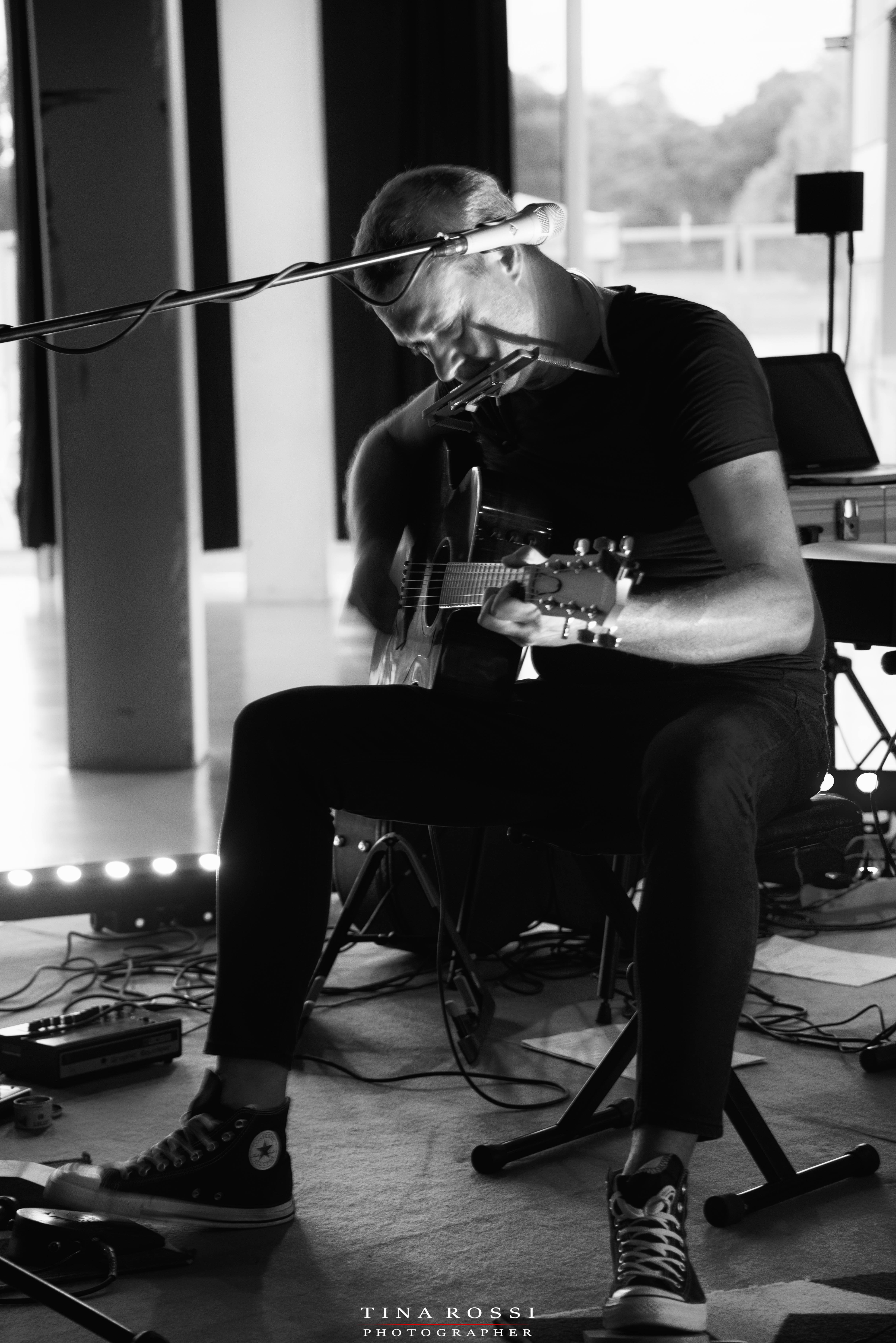 Alex D'Herin live nella foto in bianco e nero seduto davanti a un microfono con chitarra in mano