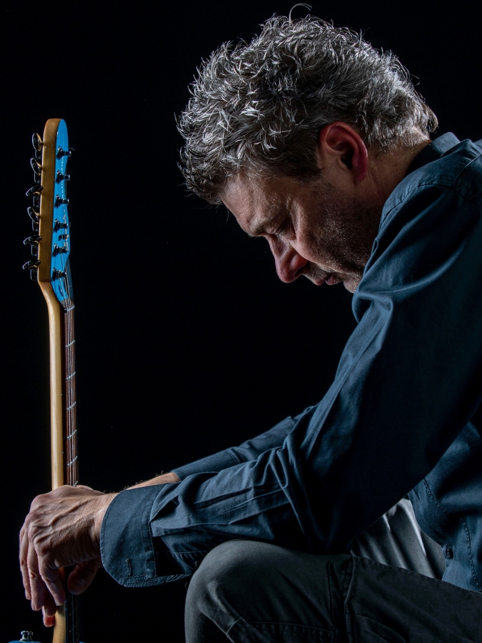 Alex D'Herin "e mi bevo un caffè" live. nella foto il cantautore torinese, di profilo, con la testa china, sedto davanti a una chitarra, che indossa camicia di jeans blu e pantaloni di tela grigia