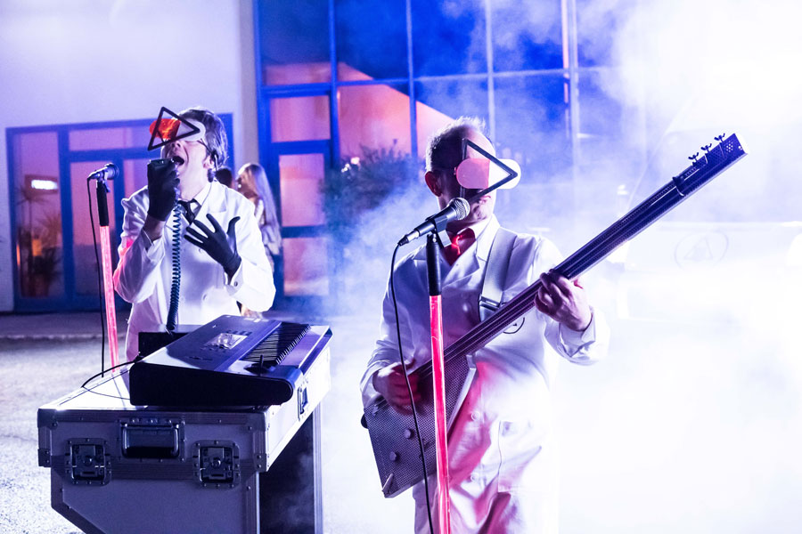 Anticorpi. Nella foto due musicisti della band, vestiti di bianco, intenti a suonare un basso e una tastiera bianchi, con strani e deformi occhiali da sole, avvolti da uno spesso fumo bianco.