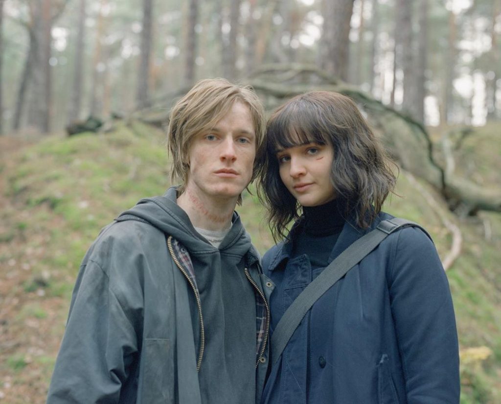 In foto Jonas e Martha giovani che guardano in camera, lui è a sinistra, lei a destra e poggia leggermente il capo su di lui. Sono vestiti con abiti neri e grigi. Sullo sfondo la foresta.