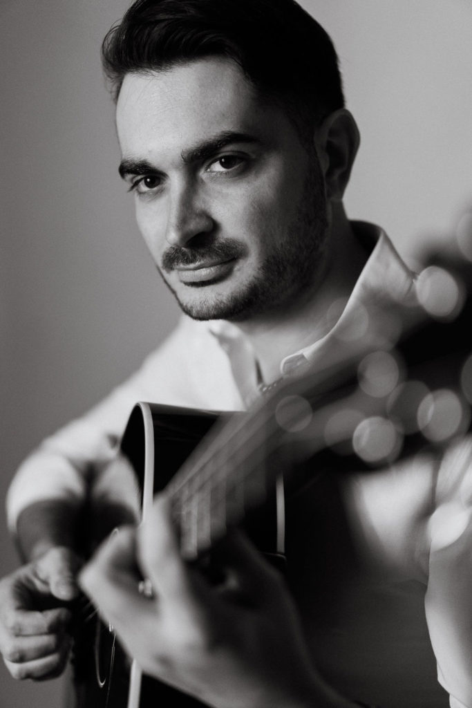 mondo di gianluca centenaro. nella fotoin bianco e nero il cantante, barba e baffi, camicia bianca che imbraccia una chitarra acustica